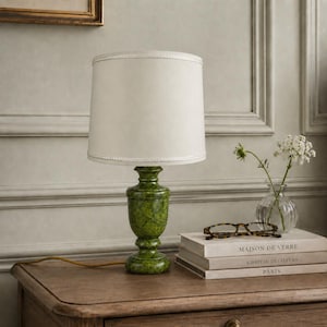 May include: A green marble table lamp with a cream-colored lampshade. The lamp sits on a wooden table next to a stack of books and a small vase of flowers. The lamp's base has a decorative, urn-like shape.