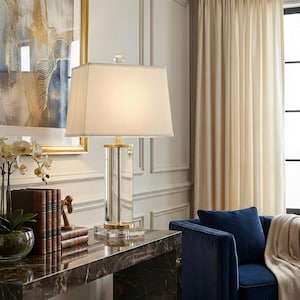 May include: A rectangular table lamp with a white lampshade and a clear, rectangular base. The base has gold-colored accents and a crystal-like finial. The lamp is on a dark marble-like table, with a painting and a blue sofa in the background.