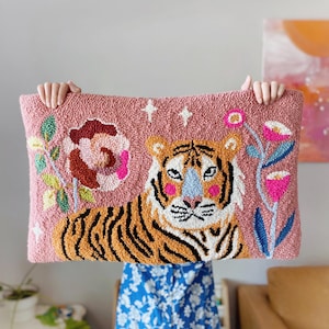 Könnte beinhalten: Ein rechteckiges, rosa Zierkissen mit einem Tigerdesign. Der Tiger hat orange und schwarze Streifen, mit rosa Wangen und einer blauen Nase. Blumen- und Sternakzente umgeben den Tiger. Das Kissen wird von zwei Händen gehalten.
