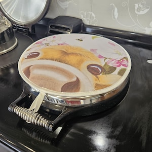 May include: A round, white stovetop cover with a brown and white cow face and pink flowers printed on it. The cover has a silver rim and a black handle with a spring.