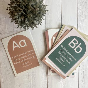 May include: A stack of inspirational cards with a neutral color palette. One card features the letter "Aa" and a quote from Proverbs 15:1. Another card displays the letter "Bb" and a quote from Ephesians 4:32. A small, green plant sits in the background.
