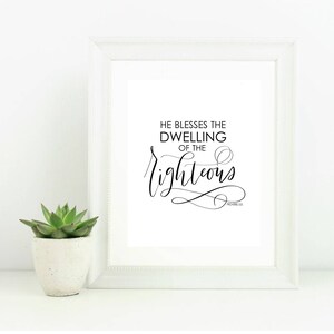 May include: A framed print with the text "HE BLESSES THE DWELLING OF THE righteous" in black script. The white frame surrounds a white background. A small succulent in a white pot sits to the left of the frame.