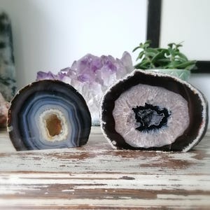 May include: Two polished agate geodes, one with blue, grey, and brown banding, the other with a white and black centre. A cluster of purple amethyst crystals is in the background. The geodes are displayed on a distressed wooden surface.
