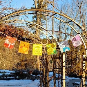 Puede incluir: Una puerta de metal oxidado con la parte superior curvada, decorada con banderas de tela de colores. La puerta está abierta y conduce a un camino nevado.