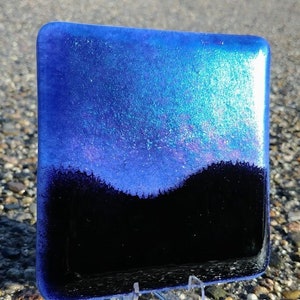 Könnte beinhalten: Eine quadratische Glasfliese mit einem blauen, schillernden Hintergrund und einer schwarzen Silhouette einer Bergkette am unteren Rand.