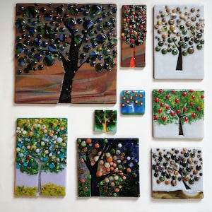 Op de afbeelding: Een verzameling van negen wandkunstwerken van glasmozaïek met verschillende boomontwerpen. De bomen zijn gemaakt van kleurrijke glasstukken en hebben een unieke, abstracte stijl. De stukken zijn gerangschikt in een rasterpatroon op een witte achtergrond.