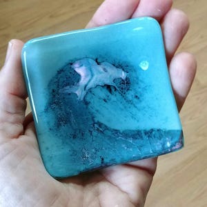 May include: A square teal blue glass coaster with a white and pink wave design.