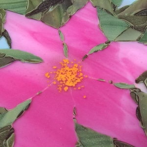 K&ouml;nnte beinhalten: Nahaufnahme einer rosa Blume mit gelber Mitte, aus Glasmosaikfliesen gefertigt. Die Blume ist von gr&uuml;nen Bl&auml;ttern umgeben.