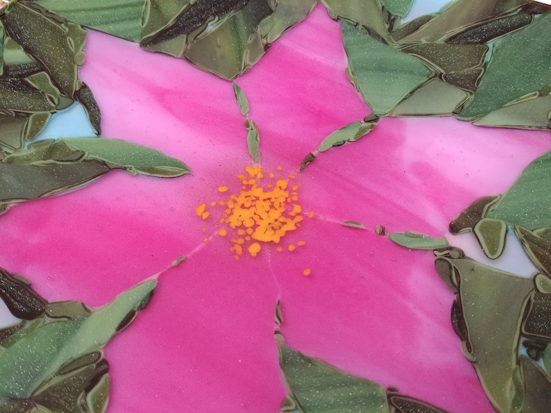 K&ouml;nnte beinhalten: Nahaufnahme einer rosa Blume mit gelber Mitte, aus Glasmosaikfliesen gefertigt. Die Blume ist von gr&uuml;nen Bl&auml;ttern umgeben.