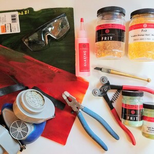 K&ouml;nnte beinhalten: Eine Sammlung von Glasmacherbedarf, darunter zwei Gl&auml;ser Frit, eine Schutzbrille, eine Atemschutzmaske, eine Zange, ein Glasschneider, eine Flasche Glaskleber und zwei Glasscheiben. Der Frit ist mit "Frit" beschriftet und die Farben sind K&uuml;rbisorange Opal und helles Aventurin.