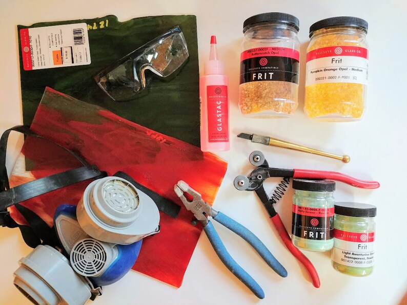 K&ouml;nnte beinhalten: Eine Sammlung von Glasmacherbedarf, darunter zwei Gl&auml;ser Frit, eine Schutzbrille, eine Atemschutzmaske, eine Zange, ein Glasschneider, eine Flasche Glaskleber und zwei Glasscheiben. Der Frit ist mit "Frit" beschriftet und die Farben sind K&uuml;rbisorange Opal und helles Aventurin.