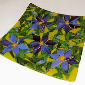 K&ouml;nnte beinhalten: Ein quadratischer Glasteller mit gelbem Hintergrund und einem Mosaikdesign aus blauen und violetten Blumen mit gelben Zentren. Die Blumen sind von gr&uuml;nen Bl&auml;ttern umgeben.