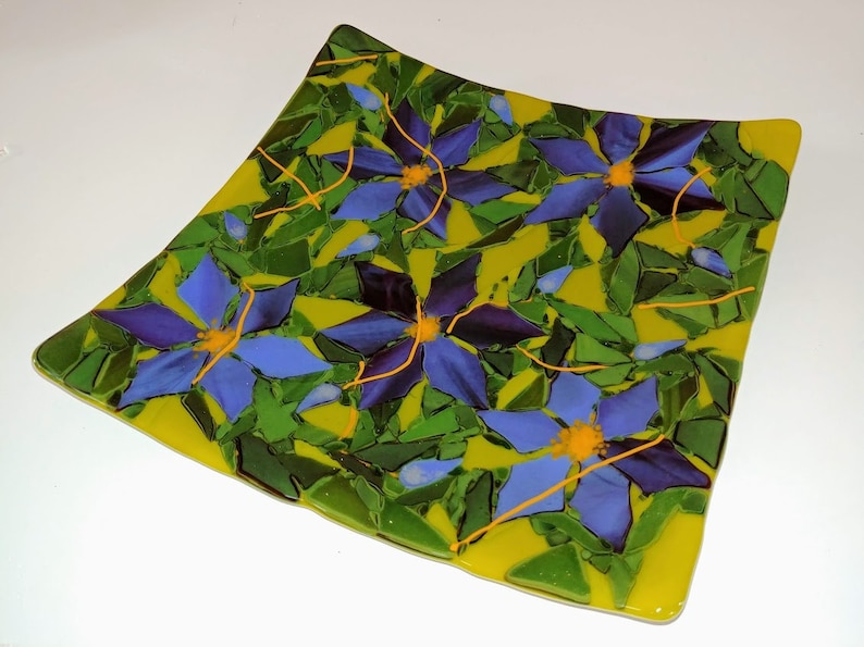 K&ouml;nnte beinhalten: Ein quadratischer Glasteller mit gelbem Hintergrund und einem Mosaikdesign aus blauen und violetten Blumen mit gelben Zentren. Die Blumen sind von gr&uuml;nen Bl&auml;ttern umgeben.
