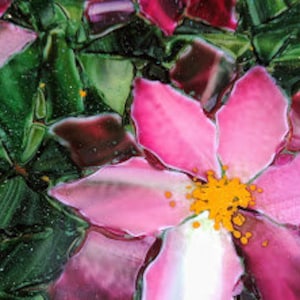 K&ouml;nnte beinhalten: Nahaufnahme einer rosa Blume mit gelber Mitte, umgeben von gr&uuml;nen Bl&auml;ttern. Die Blume ist aus Glasmosaik.