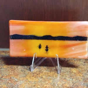 Op de afbeelding: Een rechthoekig glaskunstwerk dat een zonsondergangscène uitbeeldt. Het kunstwerk heeft oranje- en geeltinten, met een zwarte lijn voor de horizon. Twee kleine figuren zijn zichtbaar op de voorgrond. Het wordt tentoongesteld op een transparante standaard.