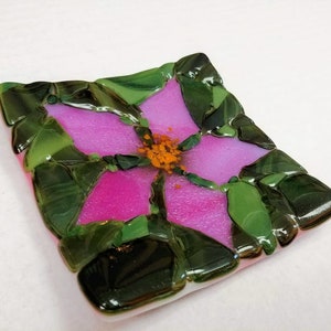 K&ouml;nnte beinhalten: Eine quadratische Glasschale mit einem rosafarbenen Blumendesign. Die Blume hat eine gelbe Mitte und gr&uuml;ne Bl&auml;tter. Die Schale ist aus verschmolzenem Glas.