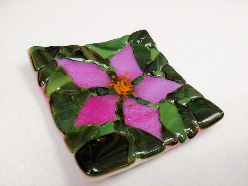 K&ouml;nnte beinhalten: Eine quadratische Glasschale mit einem rosafarbenen Blumendesign. Die Blume hat eine gelbe Mitte und gr&uuml;ne Bl&auml;tter. Die Schale ist aus verschmolzenem Glas.