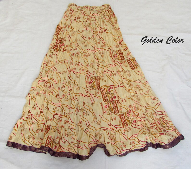 May include: A long, tiered skirt in a golden cream color featuring a repeating pattern of red and brown floral motifs and wavy lines. The skirt has an elasticized waistband and a dark purple hemline. The text Golden Color is visible.