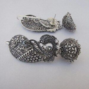 May include: Silver earrings with detailed filigree and floral patterns. The earrings have a teardrop shape with a dangling bell. The metalwork is ornate, displaying a vintage style. The earrings are approximately 6 cm in length.