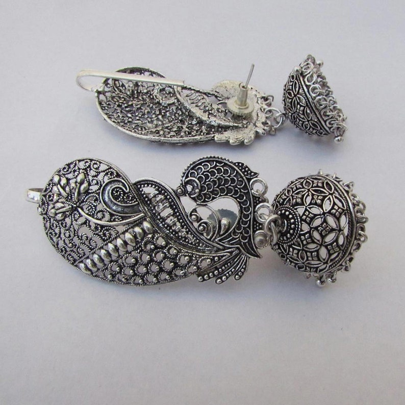 May include: Silver earrings with detailed filigree and floral patterns. The earrings have a teardrop shape with a dangling bell. The metalwork is ornate, displaying a vintage style. The earrings are approximately 6 cm in length.