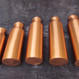 May include: Five copper-colored water bottles of varying sizes are arranged on a dark gray surface. The bottles have a cylindrical shape with a rounded base and a threaded neck. The copper finish gives them a shiny, metallic appearance.
