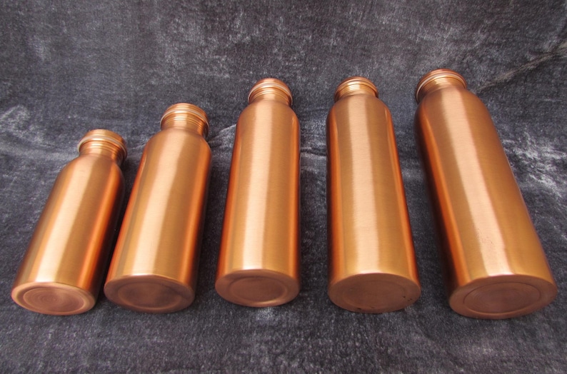 May include: Five copper-colored water bottles of varying sizes are arranged on a dark gray surface. The bottles have a cylindrical shape with a rounded base and a threaded neck. The copper finish gives them a shiny, metallic appearance.