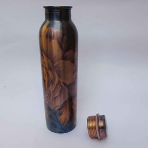 May include: A copper water bottle with a floral design in shades of brown and gold. The bottle has a matching copper cap next to it. The bottle is cylindrical and appears to be made of metal.