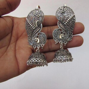 May include: A pair of silver earrings with a peacock design. The earrings have an intricate, openwork pattern and a bell-shaped base with small dangling beads. The peacock's body is accented with gold-coloured metal.