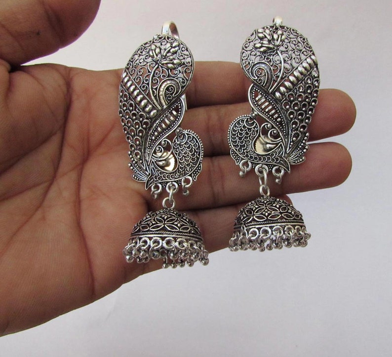 May include: A pair of silver earrings with a peacock design. The earrings have an intricate, openwork pattern and a bell-shaped base with small dangling beads. The peacock's body is accented with gold-coloured metal.