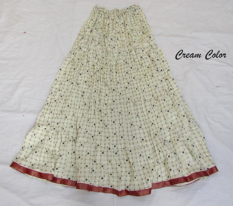 May include: A cream-colored tiered skirt featuring a repeating grid pattern with small dark circular accents. The skirt has an elasticized waistband and a wide, dark red border along the bottom hem. The text "Cream Color" is printed on the right.