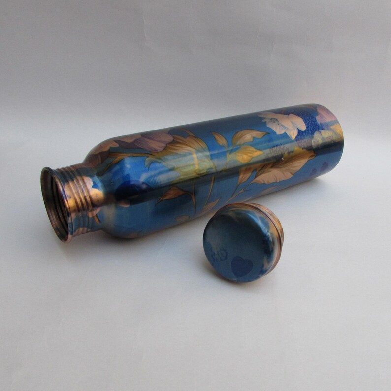 May include: A blue water bottle with a floral design in shades of purple, gold, and white. The bottle is lying on its side with the cap next to it. The bottle has a metallic finish.
