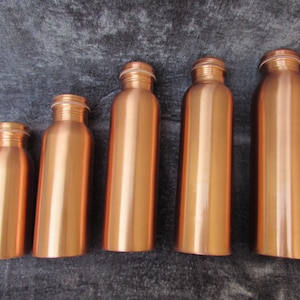 May include: Five copper-colored water bottles of varying sizes are arranged in a row. Each bottle has a cylindrical body, a narrow neck, and a screw-on cap. The bottles are made of a shiny, metallic material, reflecting the light. The background is a dark, textured surface.