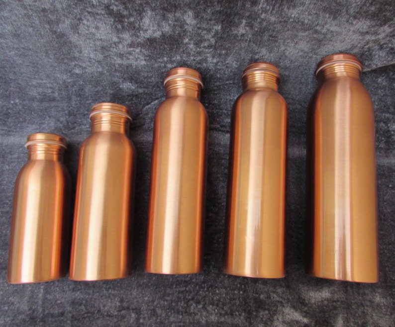 May include: Five copper-colored water bottles of varying sizes are arranged in a row. Each bottle has a cylindrical body, a narrow neck, and a screw-on cap. The bottles are made of a shiny, metallic material, reflecting the light. The background is a dark, textured surface.