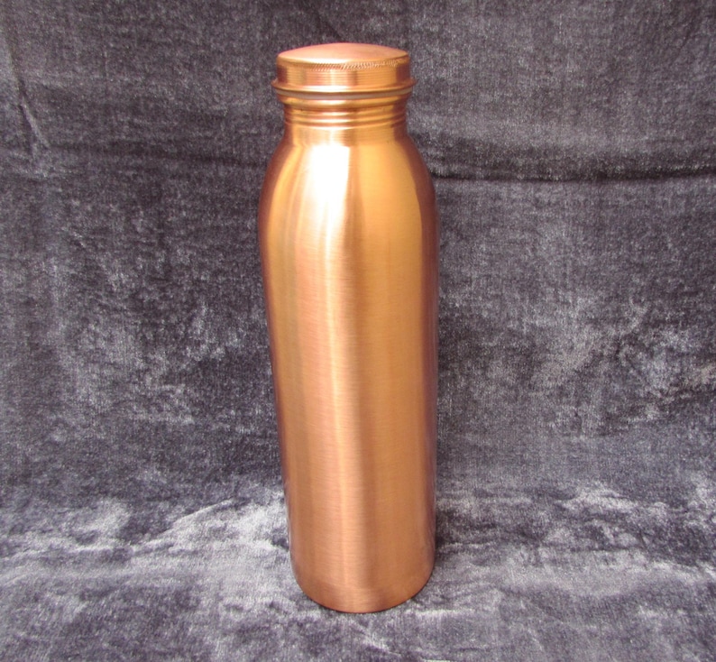 May include: A copper water bottle with a screw-on lid. The bottle has a cylindrical shape with a slightly curved body. The copper has a polished, reflective surface.