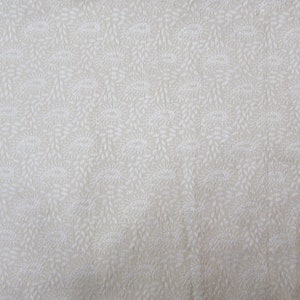 May include: A light beige fabric featuring a repeating white pattern of small, intricate paisley motifs and delicate leaf-like shapes. The design is evenly distributed across the surface, creating a subtle, textured appearance on the textile.