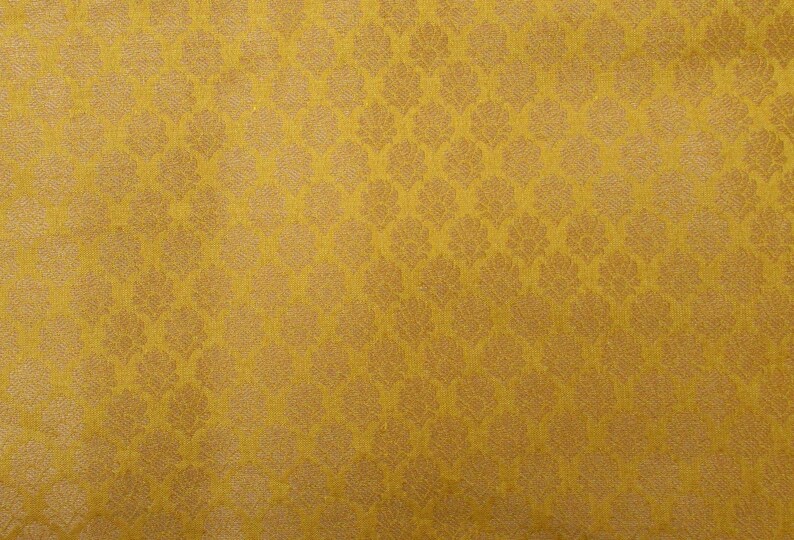 May include: A mustard yellow fabric featuring a repeating damask pattern in a lighter shade of gold. The textile displays a consistent woven texture with a symmetrical arrangement of stylized floral motifs across the entire surface area.