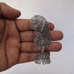 May include: Silver-coloured peacock-shaped earring with detailed filigree work. The earring has a stylised peacock design with a bell-shaped dangle. The metal has a slightly oxidised finish, enhancing the details. The earring is held in a hand.
