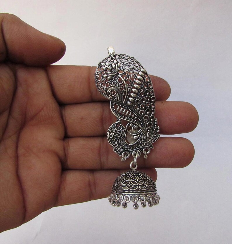 May include: Silver-coloured peacock-shaped earring with detailed filigree work. The earring has a stylised peacock design with a bell-shaped dangle. The metal has a slightly oxidised finish, enhancing the details. The earring is held in a hand.