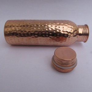 May include: A cylindrical copper water bottle with a hammered texture finish. The bottle is positioned horizontally with its matching screw-top lid removed and placed in front, revealing a white silicone seal ring on the lid's threaded neck.
