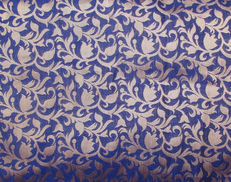 May include: A deep blue fabric featuring a repeating woven pattern of light beige floral and leaf motifs. The design consists of stylized tulip-like shapes and swirling vines that create a dense, intricate texture across the entire surface.