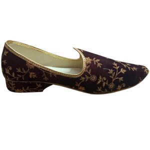 May include: A dark maroon slip-on shoe featuring an intricate gold floral embroidery pattern across the upper. The shoe has a low block heel, a pointed toe, and a metallic gold trim lining the collar and opening of the footwear.