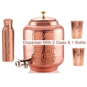 Könnte beinhalten: Ein Wasserspender aus Kupfer mit gehämmertem Muster, einem Metallhahn und einem Deckel mit kleinem Knauf. Das Set enthält eine passende gehämmerte Kupferflasche und zwei gehämmerte Kupferbecher. Der Text Dispenser With 2 Glass & 1 Bottle ist sichtbar.