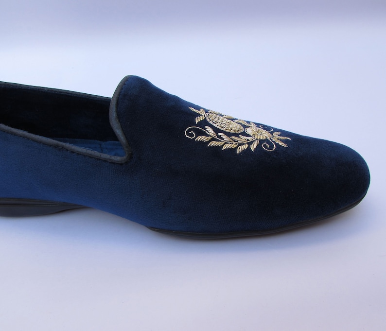 Pode incluir: Um mocassim de veludo azul profundo com um motivo bot&acirc;nico bordado a fio dourado na parte superior. O sapato tem um acabamento escuro ao longo da abertura e uma sola preta plana. O design apresenta detalhes de costura floral e foliar.