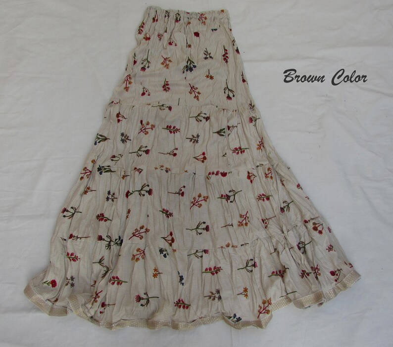 May include: A tiered maxi skirt in a light beige fabric with a fine brown pinstripe pattern and a scattered floral print in red, orange, and blue. The skirt features an elasticized waistband and a hem finished with a light brown trim. Text reads Brown Color.