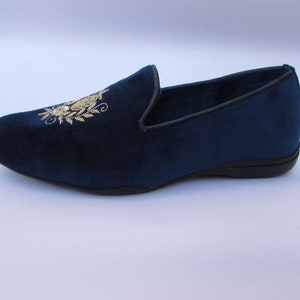 Pode incluir: Um mocassim de veludo azul-marinho com um motivo floral bordado a fio dourado no peito do p&eacute;. O sapato apresenta uma biqueira arredondada, uma silhueta de perfil baixo e uma sola preta com um rebordo fino preto ao longo da abertura do sapato.
