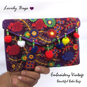 Può includere: Una pochette viola ricamata con motivi floreali colorati e pompon pendenti. La pochette presenta ricami intricati in rosso, blu, giallo e verde, con dettagli di perline dorate. Il testo sulla borsa recita "Lovely Bags" e "Embroidery Vintage Beautiful Boho Bags."