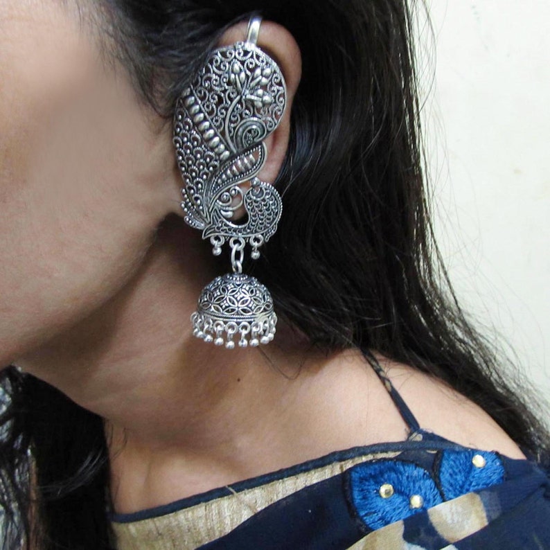 May include: Silver-coloured ear cuff and earring set. The ear cuff features a peacock design with intricate detailing, and the earring has a bell shape with dangling beads. The jewellery is worn on the ear.