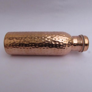 May include: A cylindrical copper water bottle featuring a hammered texture across its body. The bottle has a smooth, narrow neck with horizontal ridges and a matching screw-top cap. The metallic surface reflects light, showing a warm, reddish-brown tone.