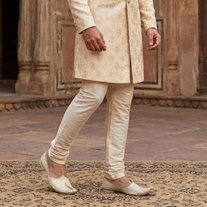 May include: A cream-colored sherwani jacket featuring a subtle gold floral pattern, paired with matching cream-colored slim-fit trousers. The outfit is completed with cream-colored slip-on shoes that have a textured, woven-like surface finish.