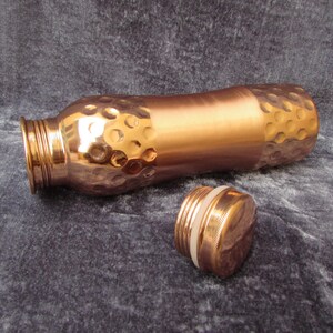 May include: A copper water bottle with a hammered texture on the top and bottom sections. The bottle is lying on its side with the cap unscrewed and next to it. The bottle has a smooth, polished finish in the middle section.